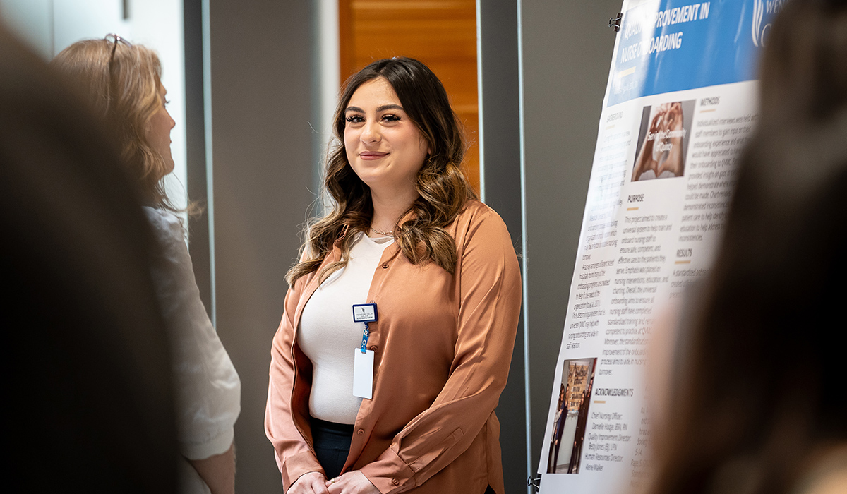 An RN to BSN graduate stands next to her research poster and smiles at the camera.