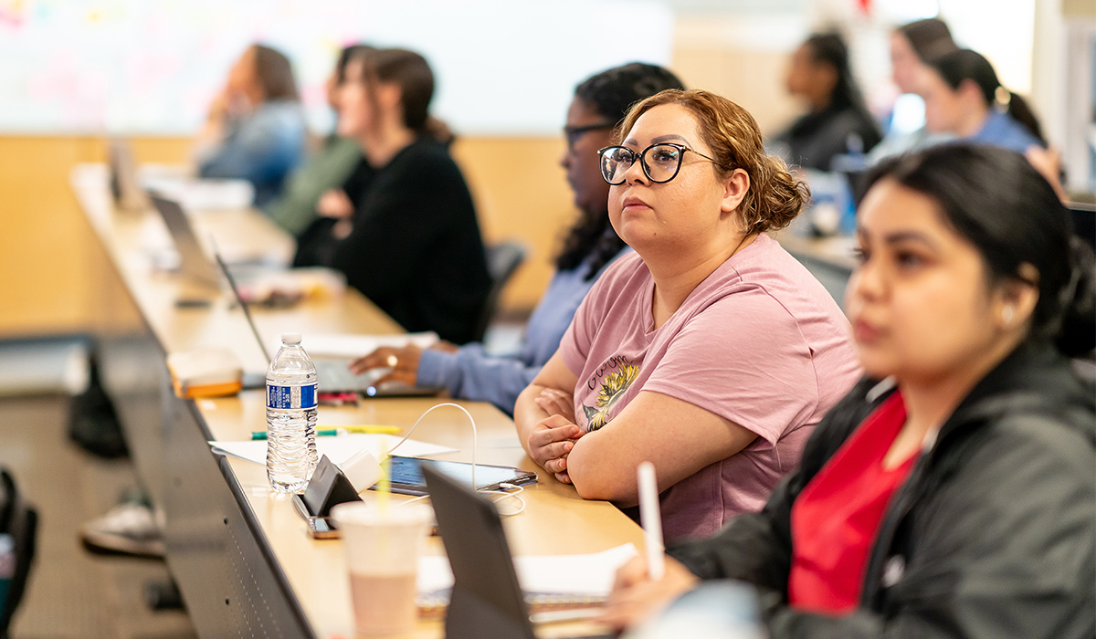 A group of students sit at long tables and look toward the front of the classroom.