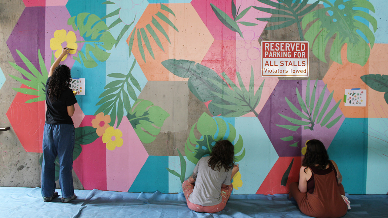 Three students paint a plant and multi-colored mural on a concrete wall.