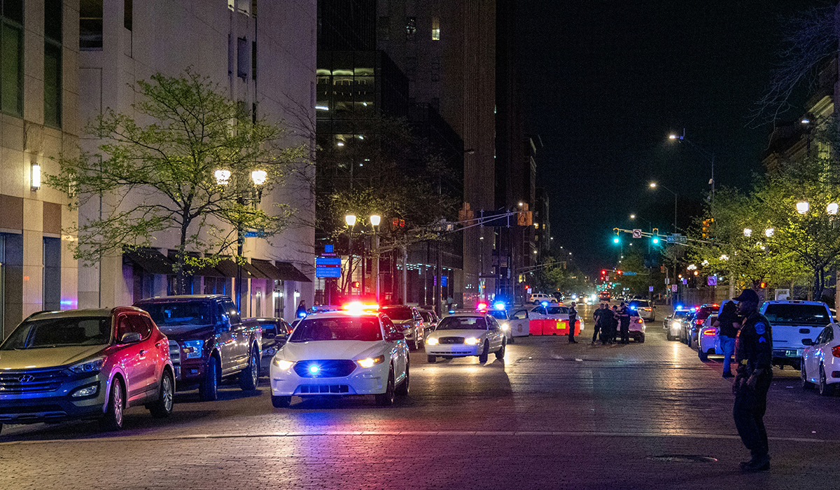 Multiple police cars with blue and red lights are parked on a dark street, with an officer crossing the street.