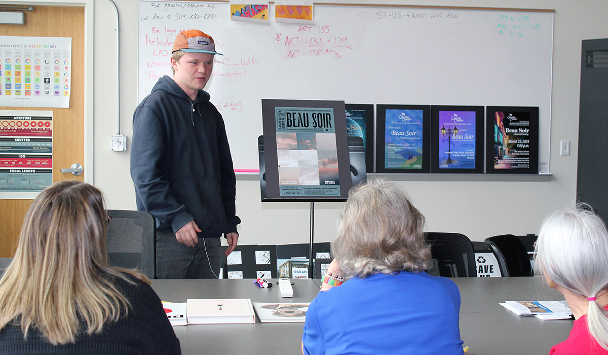 A graphic design student stand next to a poster he created while three women review it.