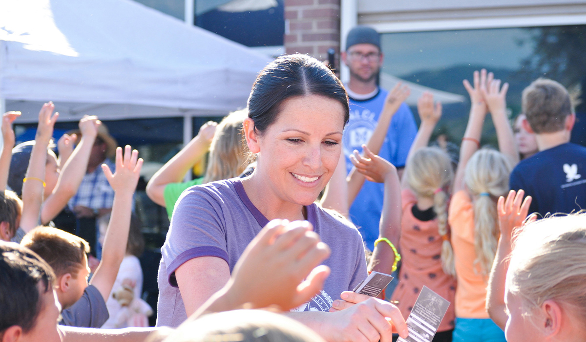 A woman stands among a group of children with their hands raised and gathers pieces of paper.