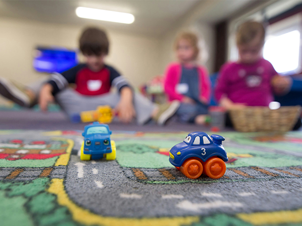 In the foreground are two toy cars on a floor mat that looks like a road; in the background are three small children.