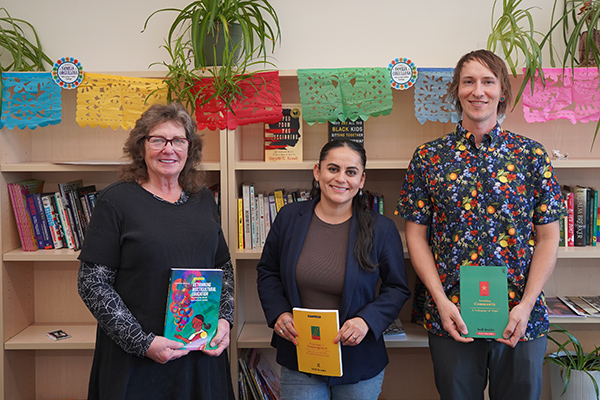 Barbara Oldham stands in front of a bookcase with El Corazon Director Maria Morales-Sanchez and Professor Saxon Spillman